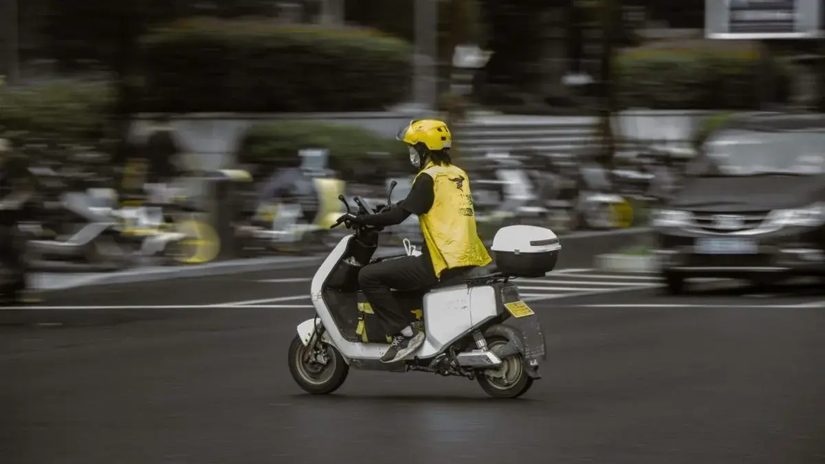 A delivery rider on a scooter, motion-blurred, weaving through daytime city traffic