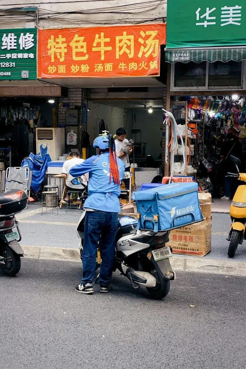 A delivery rider standing at a street-side beef noodle shop counter, insulated delivery bag on the scooter behind him