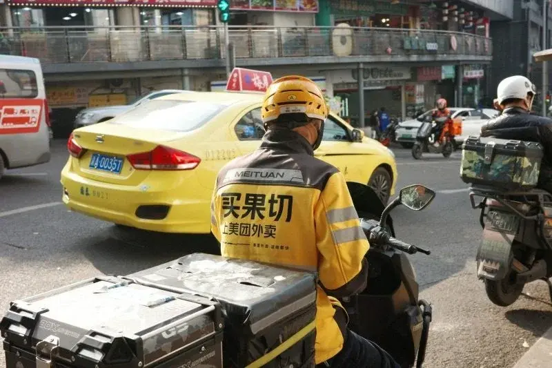 A Meituan delivery rider in a yellow jacket printed with a slogan about fast fruit-cutting delivery, waiting at a city intersection