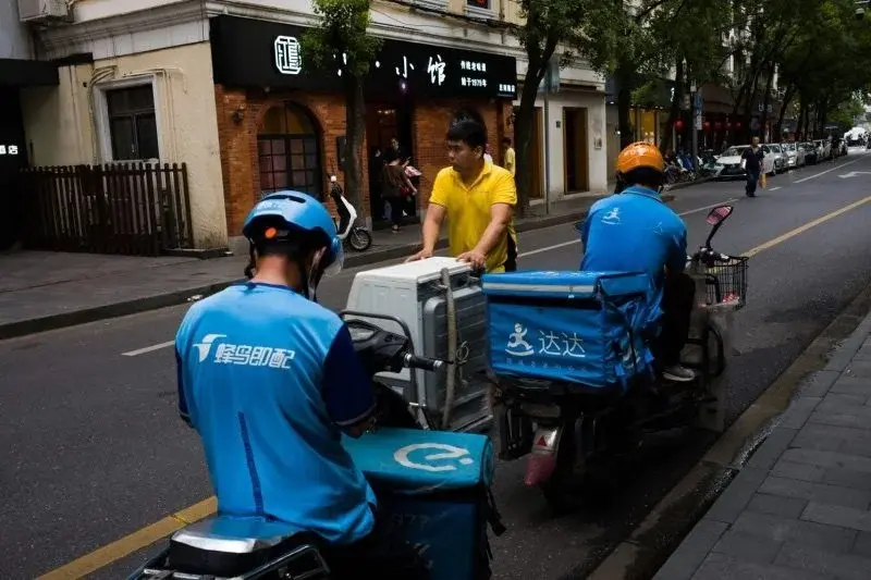 Three delivery riders from different platforms — Ele.me, Dada, and a third courier service — pausing on the same city street