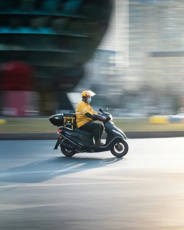 A delivery rider in a yellow uniform, motion-blurred while riding past a building, an insulated delivery bag mounted behind the seat