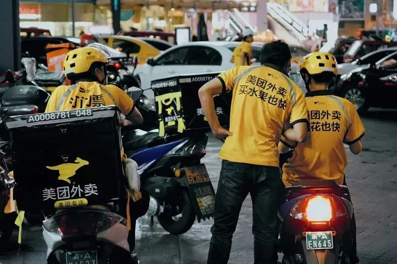 Three Meituan delivery riders in yellow uniforms printed with playful marketing slogans about fast delivery of fruit and flowers