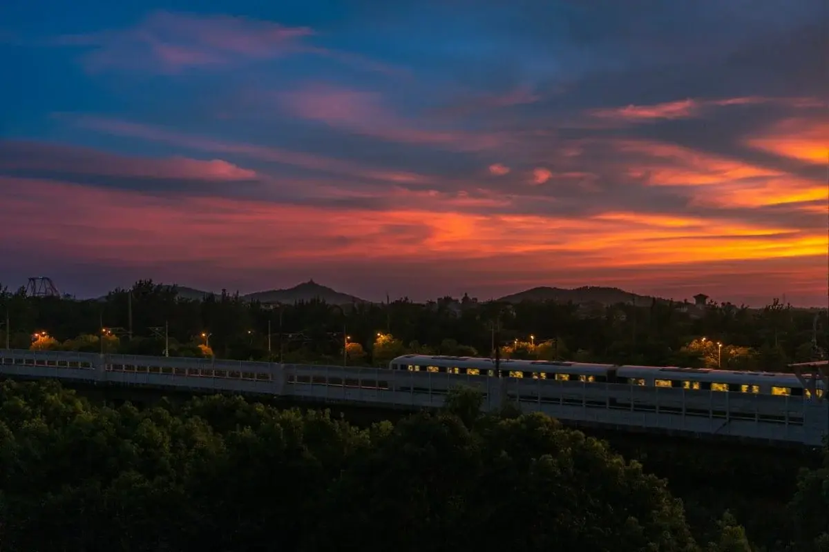 A high-speed train crossing an elevated viaduct at sunset with a dramatic orange sky