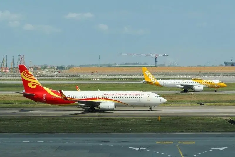 A Hainan Airlines plane and a Scoot plane taxiing on a runway, with construction cranes visible in the background