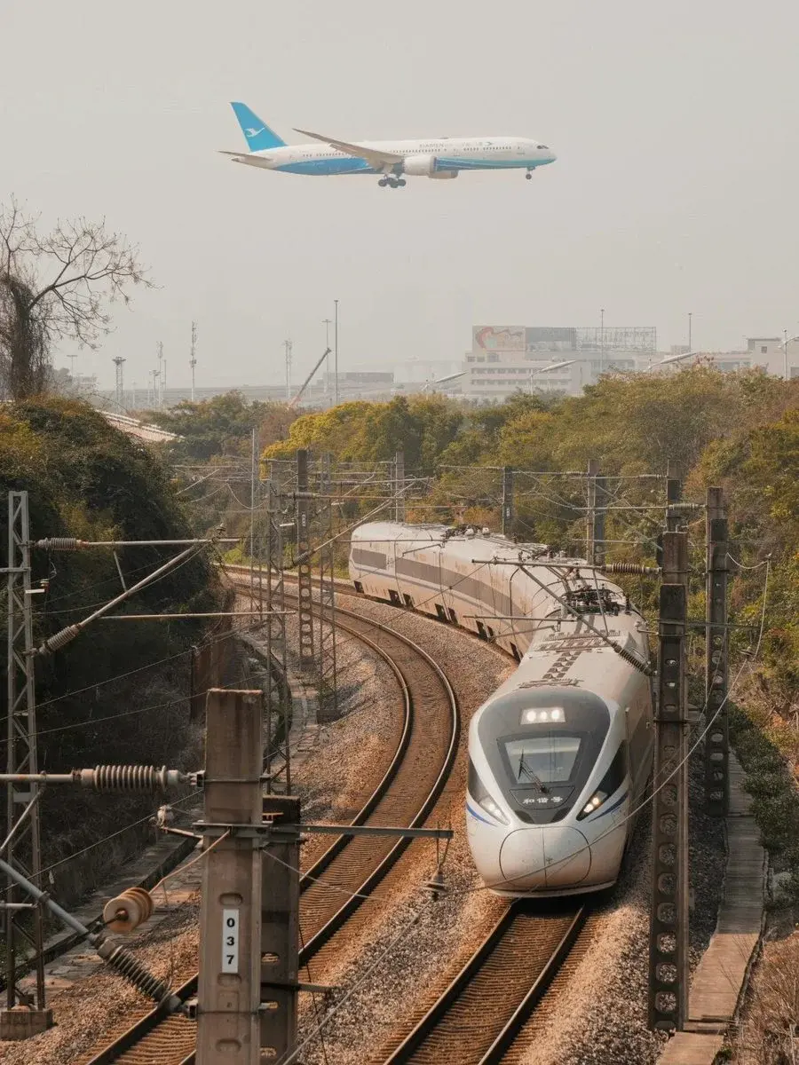A Xiamen Airlines plane descending over a curving high-speed rail line with a train passing below