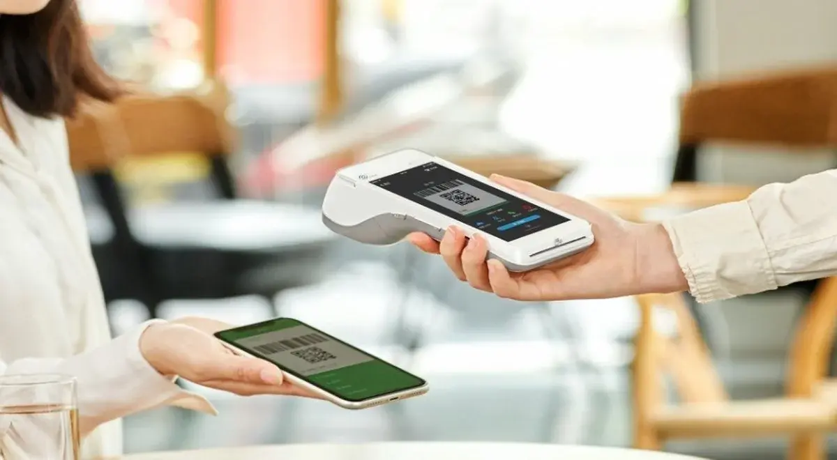Close crop of a payment terminal displaying a QR code alongside a customer's phone showing a scan-to-pay screen