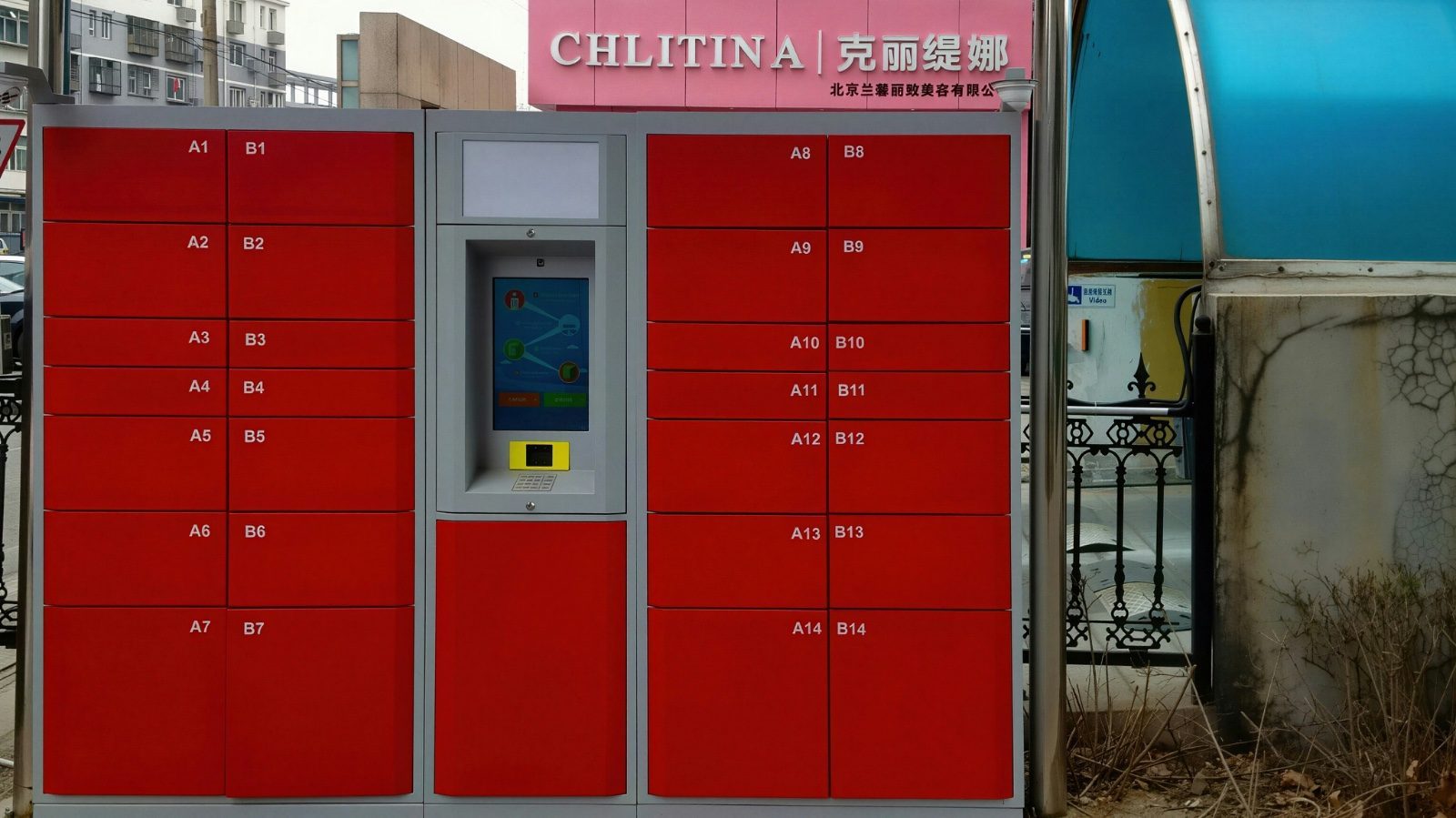 A wall of red smart parcel lockers on a city sidewalk, with a touchscreen kiosk for pickup