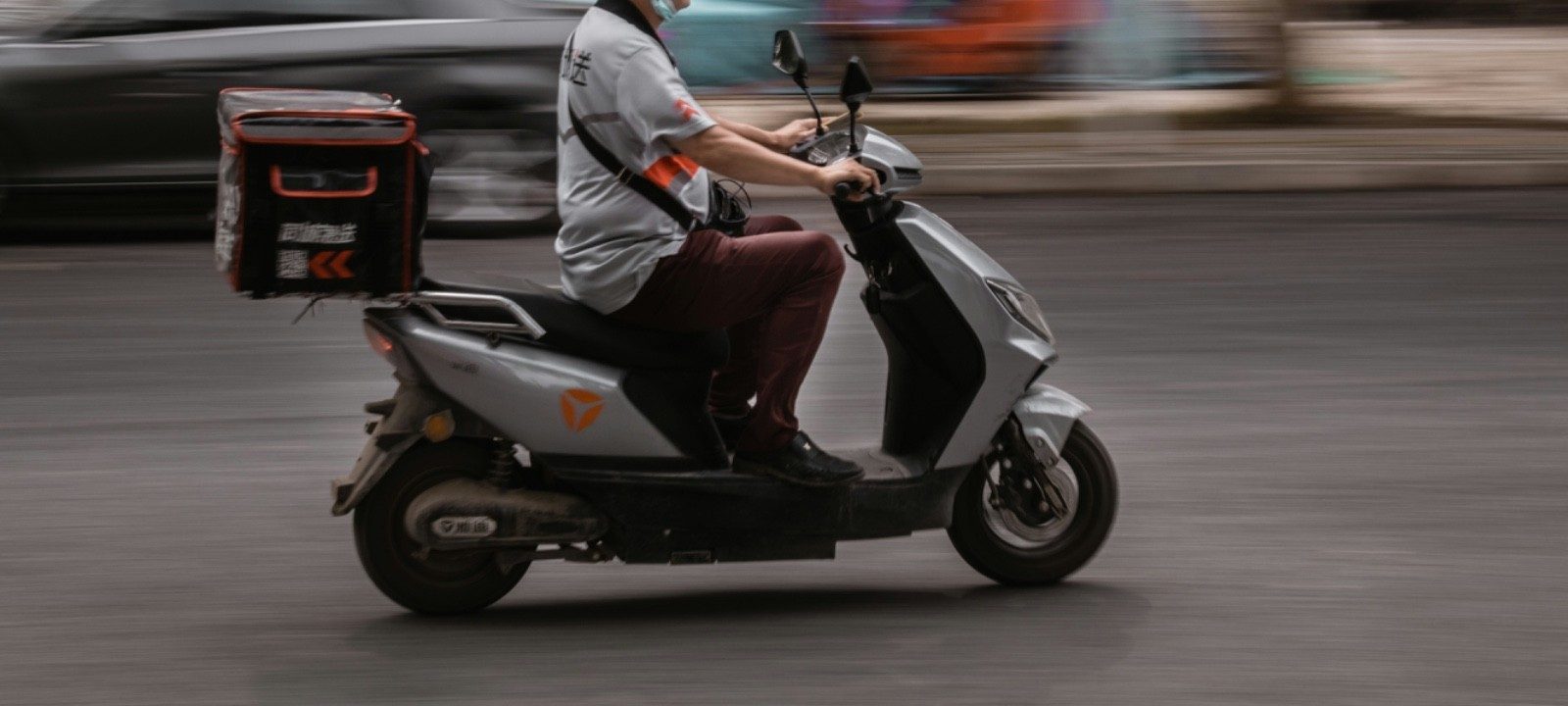 A delivery scooter in motion, insulated delivery box mounted on the back, motion-blurred street in the background