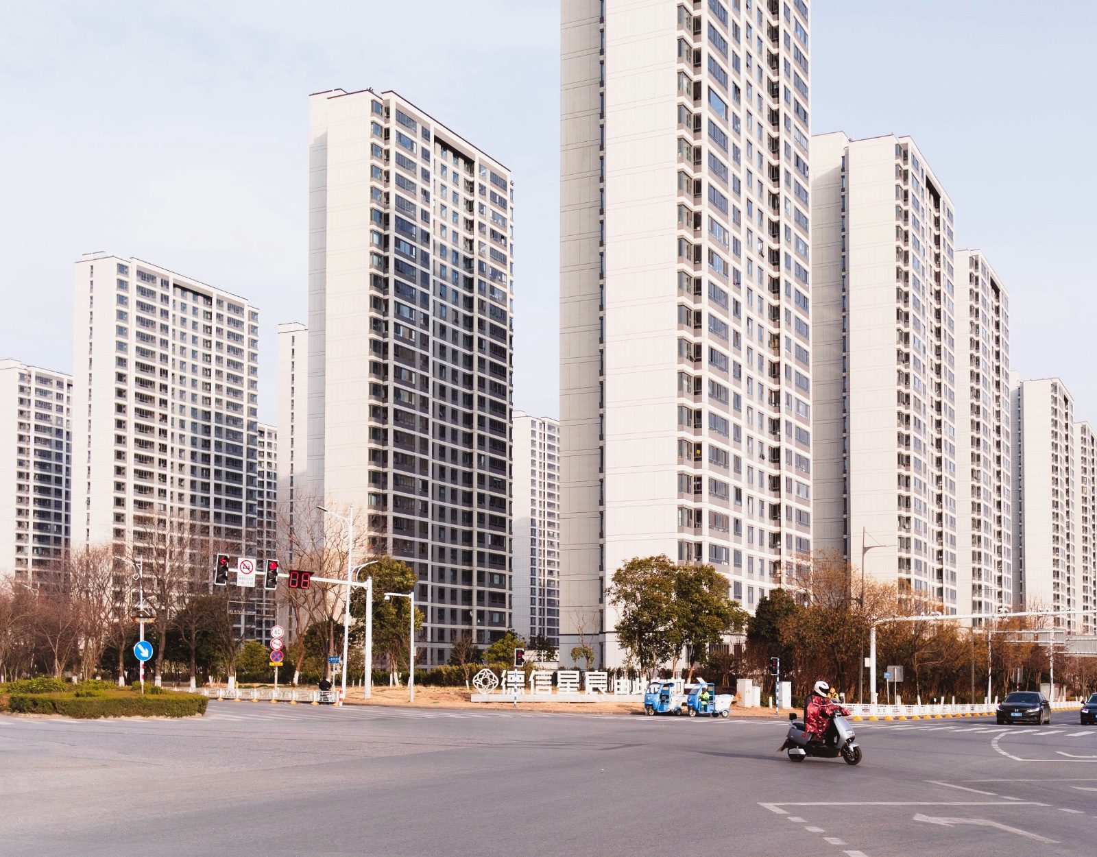 A row of newer high-rise apartment towers in Nanjing at a wide intersection, with an electric scooter crossing in the foreground