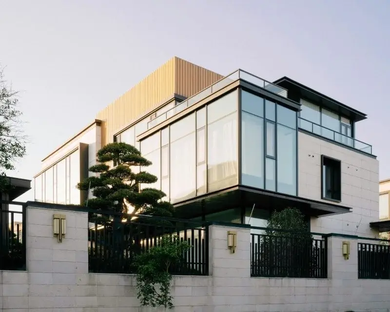 A modern glass-fronted villa in Nantong with a landscaped garden and dark metal fencing, photographed at dusk