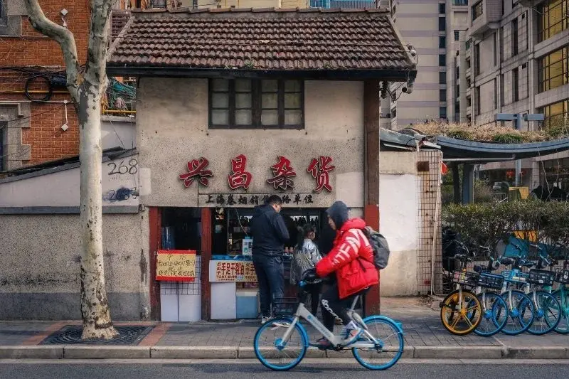 A shared bike rider passing a small shop in a Shanghai lane, with more shared bikes parked along the curb