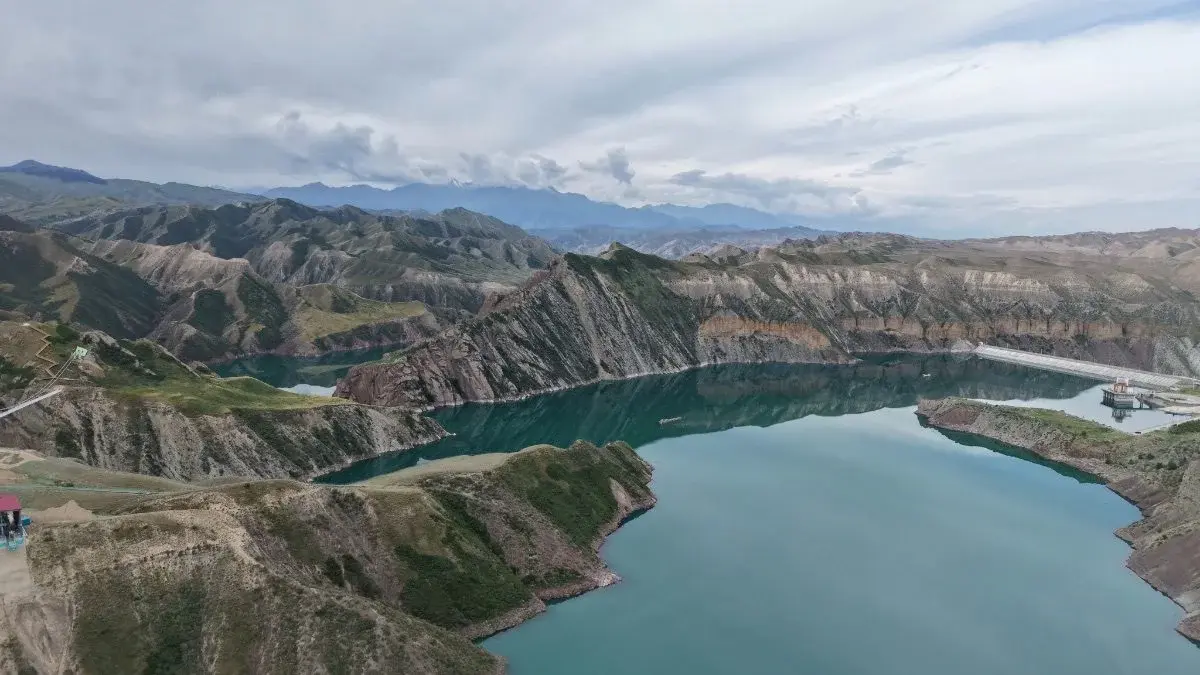 A turquoise reservoir winding through dry, terraced mountains under a cloudy sky