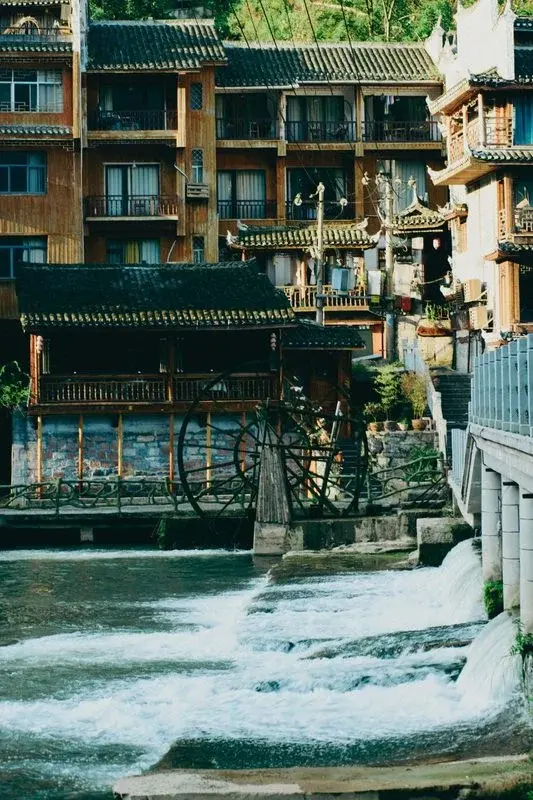 A traditional wooden waterwheel beside a river in an old riverside town, with wooden buildings along the bank