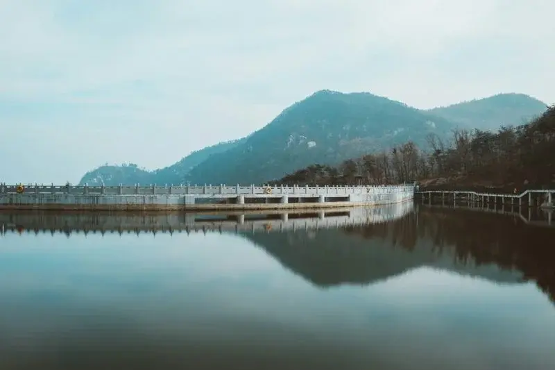 A calm reservoir with a stone balustrade walkway, reflecting misty mountains